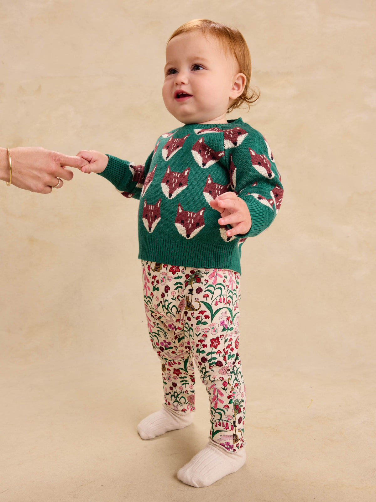 A toddler standing and holding an adult's hand, wearing the Family Fox Sweater - Foxwood Fair Isle by Tea Collection in emerald green with red-and-white fox faces across the chest and sleeves, paired with patterned floral leggings, against a neutral backdrop.