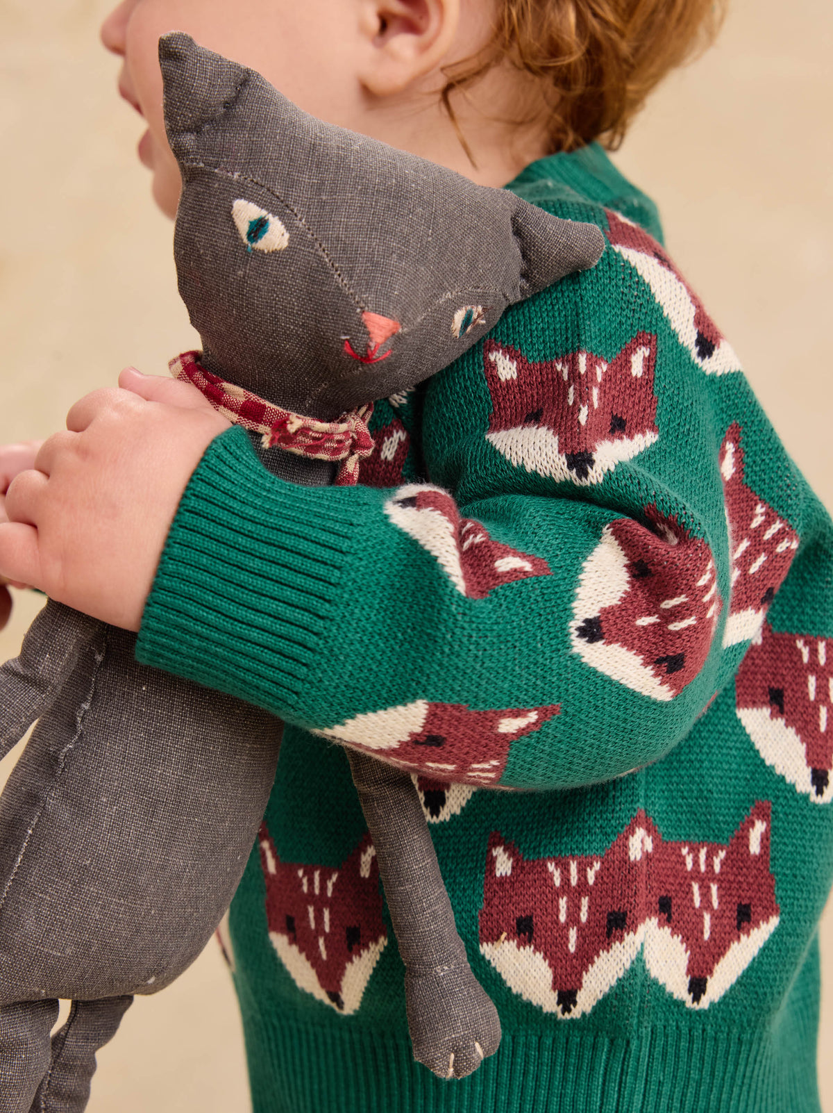 A close-up view of a child wearing the Family Fox Sweater - Foxwood Fair Isle by Tea Collection in emerald green with red-and-white fox faces across the sleeves, holding a gray stuffed fox plush toy.