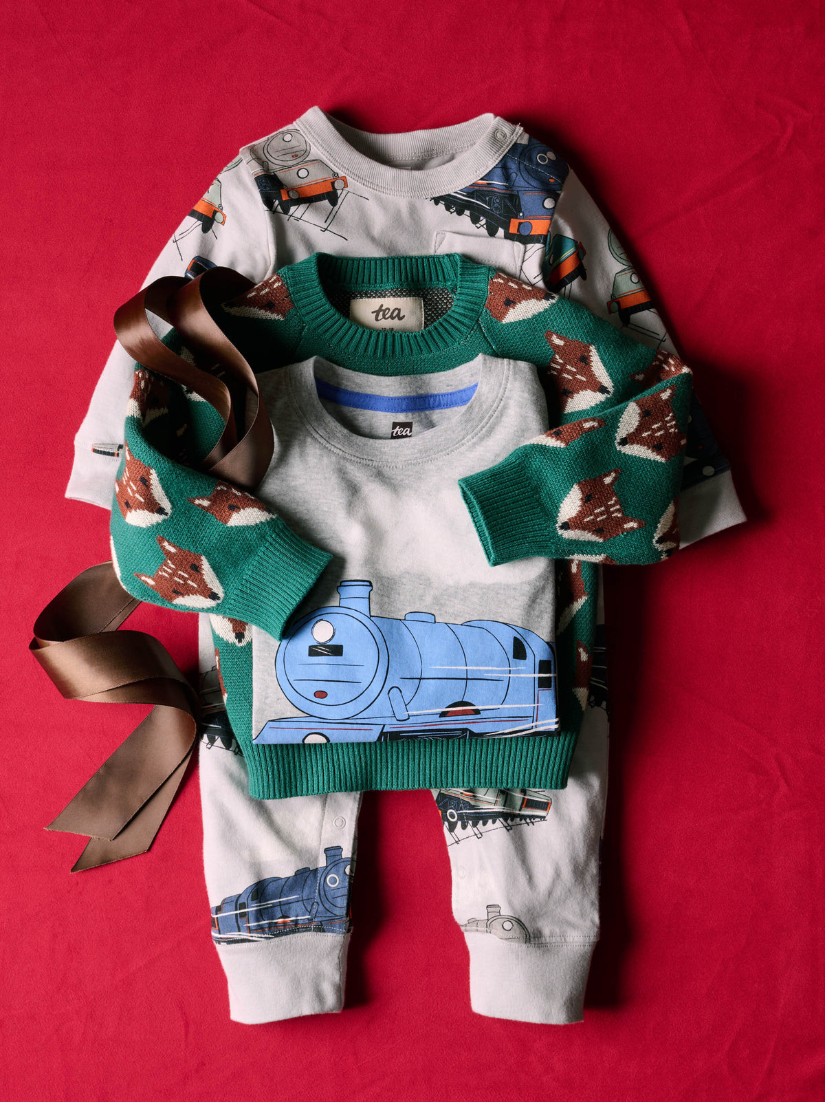 A flat lay view of a folded Family Fox Sweater - Foxwood Fair Isle by Tea Collection in emerald green with red-and-white fox faces, stacked with other children's clothing items and tied with a brown ribbon, on a red surface.