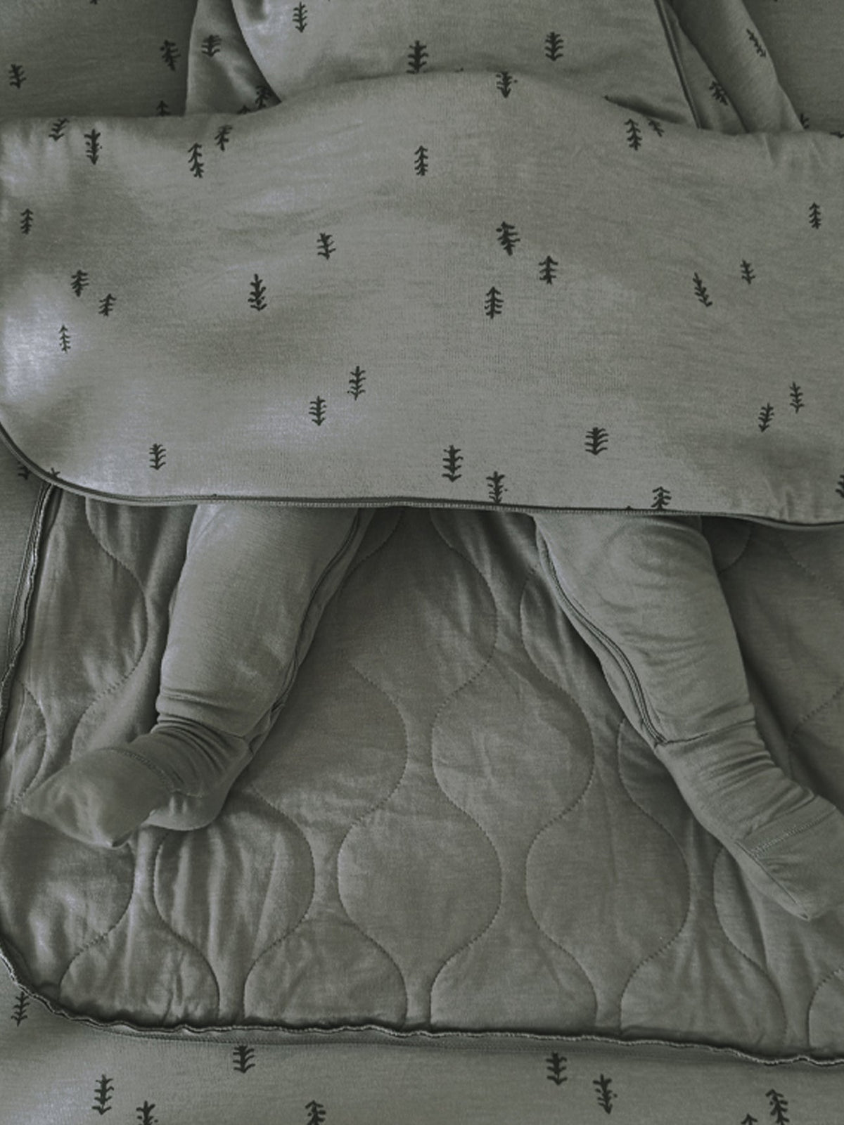 A close-up of a baby sleeping in a matching sage green quilted blanket and footed sleeper set with a small embroidered tree pattern.