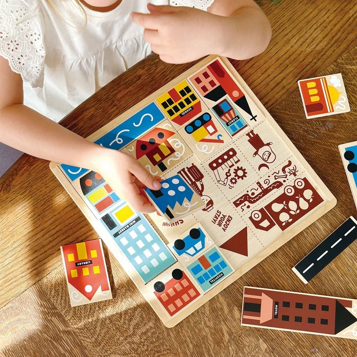 A young girl with blonde hair tied back sits at a wooden table, smiling as she plays with the Mentari Wooden Town Puzzle, holding a puzzle piece in her hand while assembling the colorful town scene.