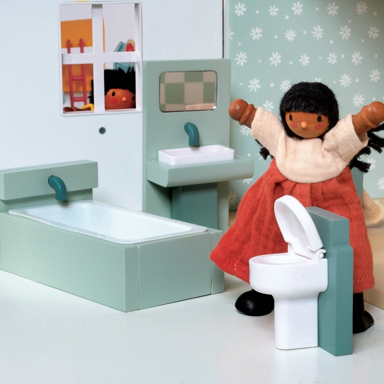 A lifestyle product photo of the Wooden Bathroom Furniture set by Mentari set up in a miniature dollhouse bathroom, featuring a doll with dark hair and a red dress standing near the toilet and sink.