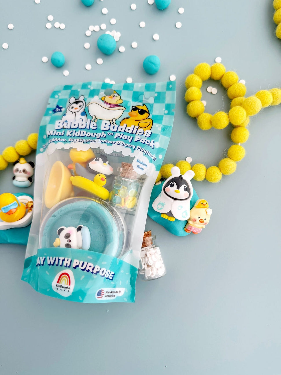 Bubble Buddies Mini KidDough Play Pack toy set in a clear plastic bag with a teal and white checkered label, displayed on a light blue-gray surface with yellow pom-pom beads and small decorative figurines around it.