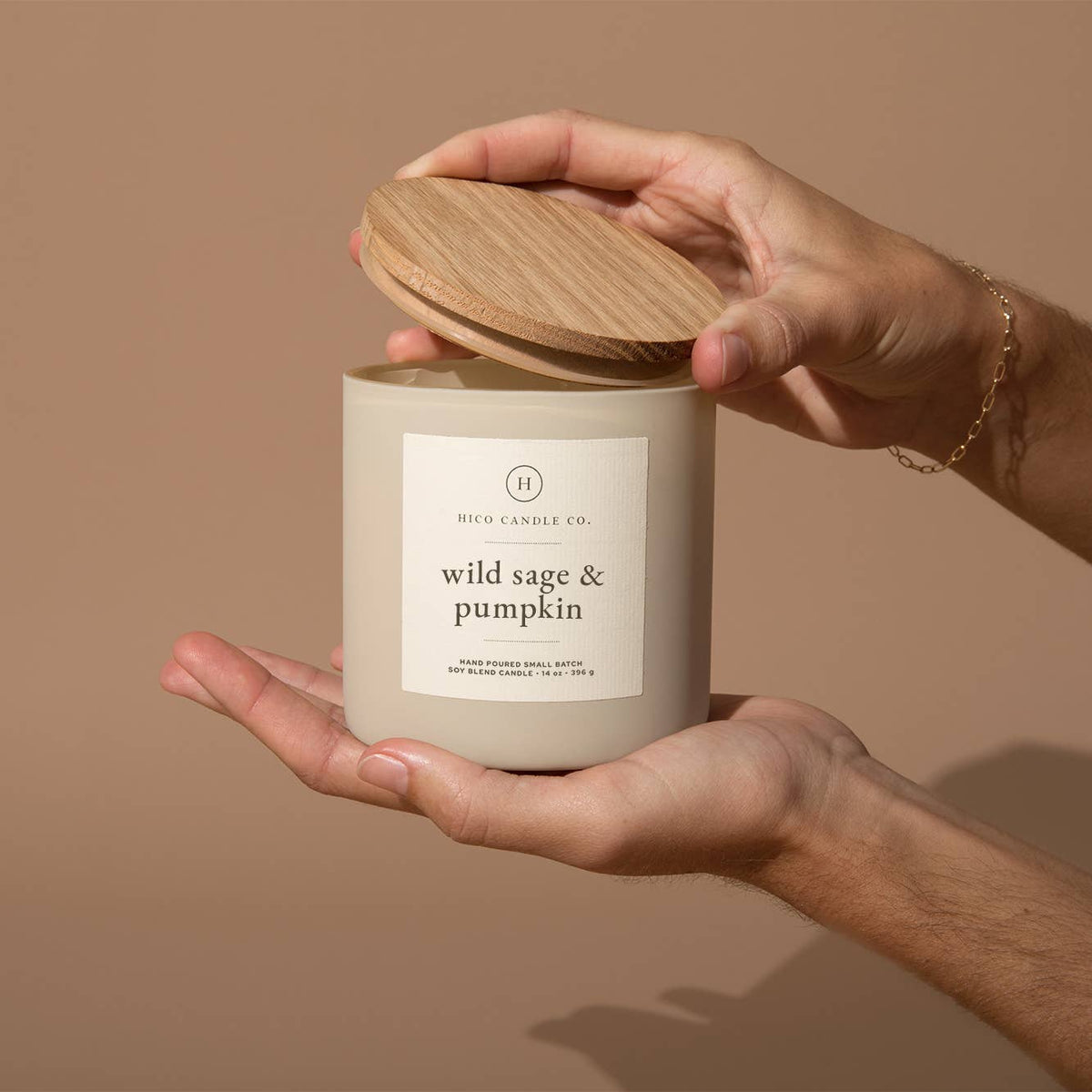 Hand holding a candle labeled 'wild sage & pumpkin' with a beige background