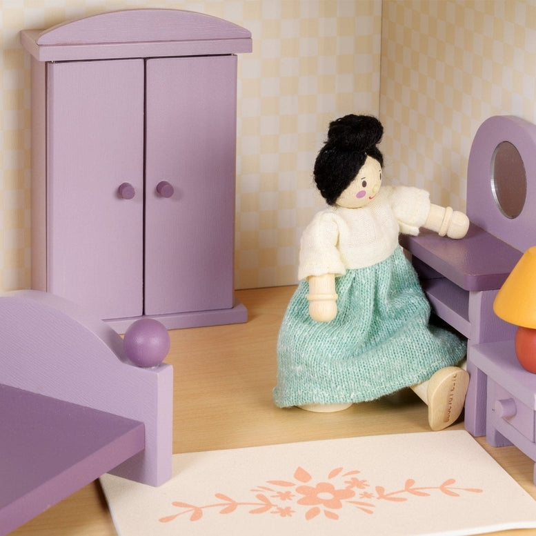 A dollhouse bedroom scene featuring a wooden doll in a green dress sitting on the edge of a double bed with a purple frame and bedding. Against the back wall stands a purple wooden wardrobe with closed doors, flanked by a dressing table with an oval mirror and a matching stool. Next to the bed is a bedside table topped with a lamp featuring an orange base and a yellow shade, and a small floral-patterned rug rests on the floor in the foreground.