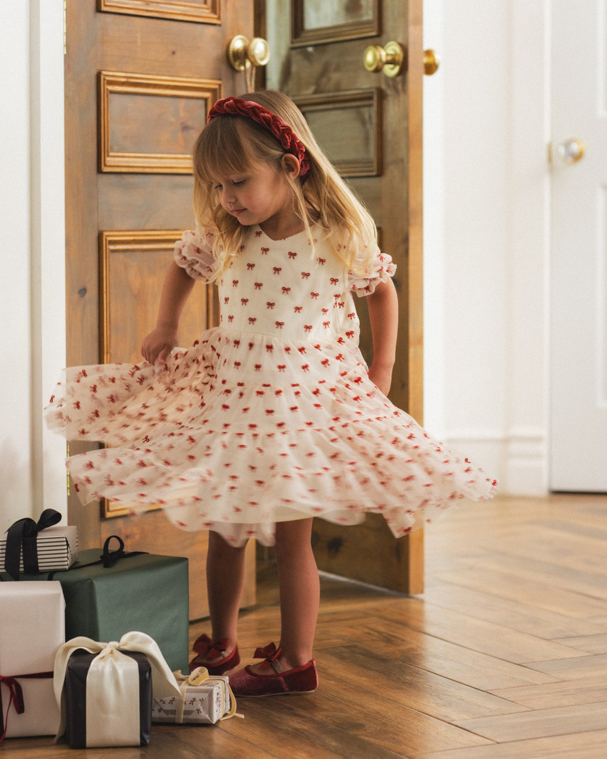 A young girl with blonde hair and a red headband twirls in a white tulle dress with red embroidered stars, standing in a doorway with wooden floors and gift boxes at her side.