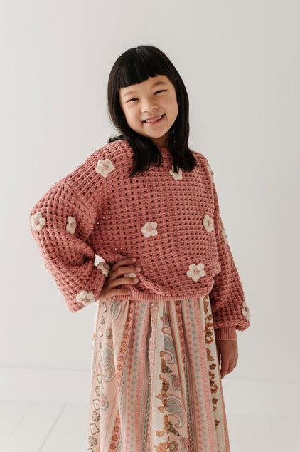A young girl with black hair smiling, wearing a dusty pink crocheted sweater with white floral accents and a long patterned skirt, standing against a plain white studio background.