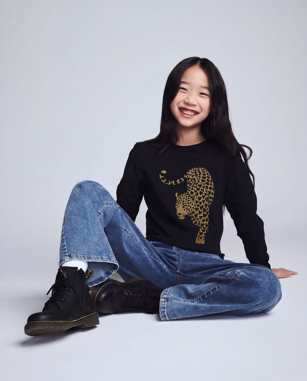 Person wearing a black sweater with a leopard print design, blue jeans, and black boots on a light gray background