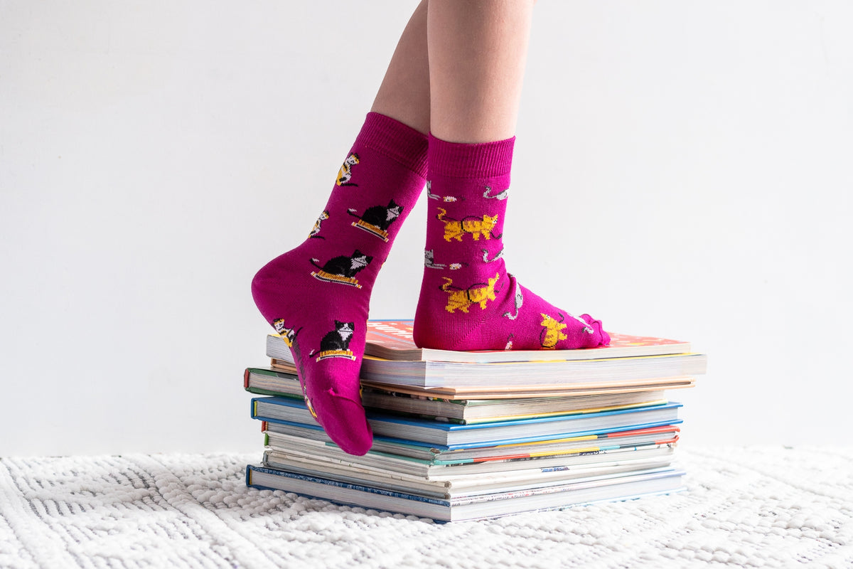 A person's legs wearing bright pink crew socks with a playful cat and dog pattern, standing on a stack of books on a white textured blanket against a white background.