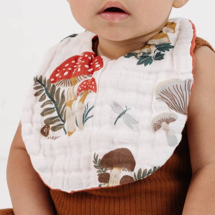 A close-up of a baby wearing a white muslin bib with a colorful printed design of red and brown mushrooms, green leaves, and a dragonfly, paired with a brown ribbed knit top.