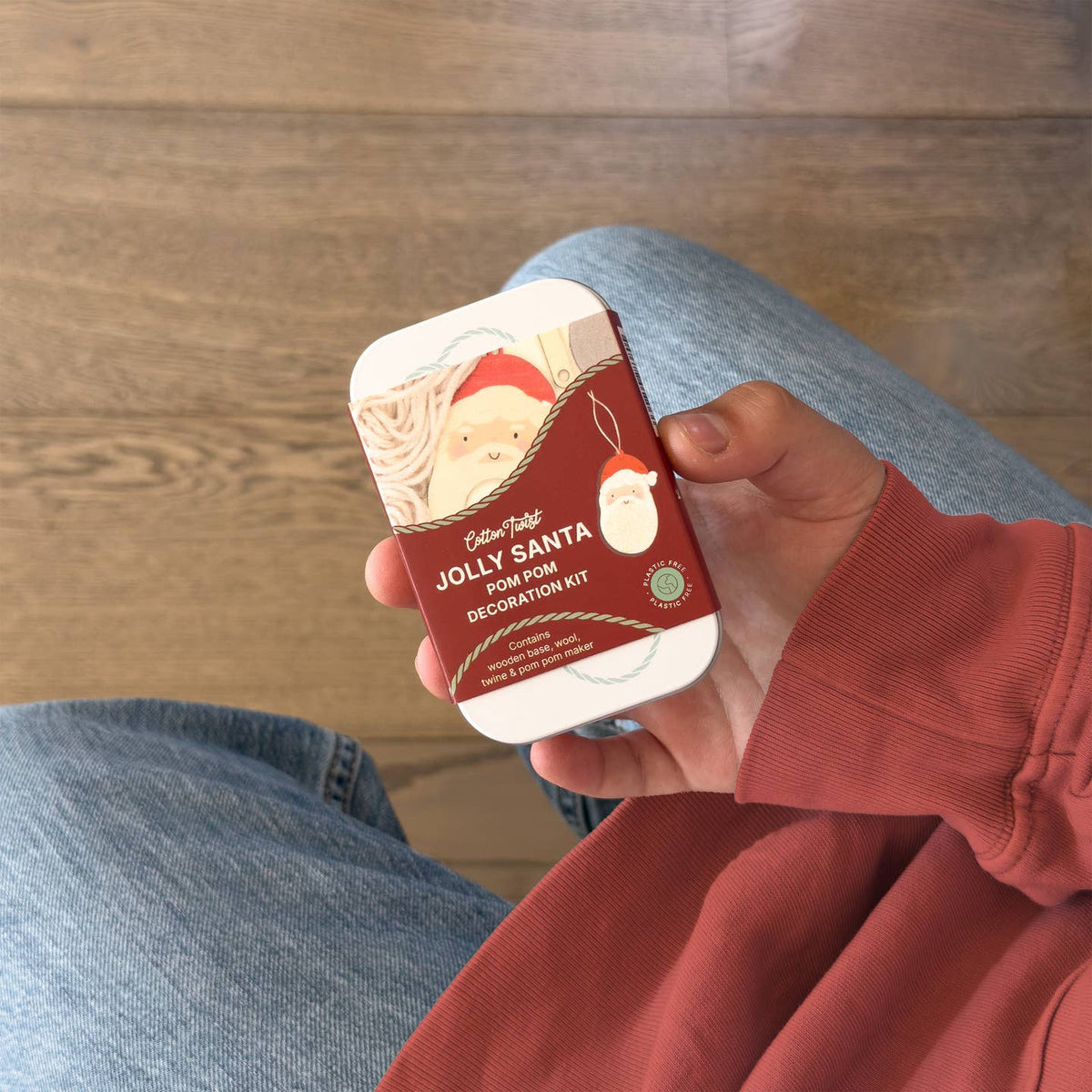A person in a rust-colored sweatshirt and blue jeans holds a small metal tin labeled 'Jolly Santa Fish Hook' and 'Decorative Kit' against a wooden floor background.