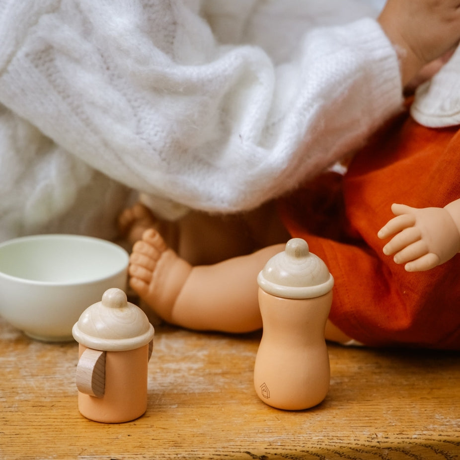 Two small wooden toy bottles with beige nipples and lids, displayed on a rustic wooden table next to a white bowl, with a person in a white sweater holding a doll in the background.