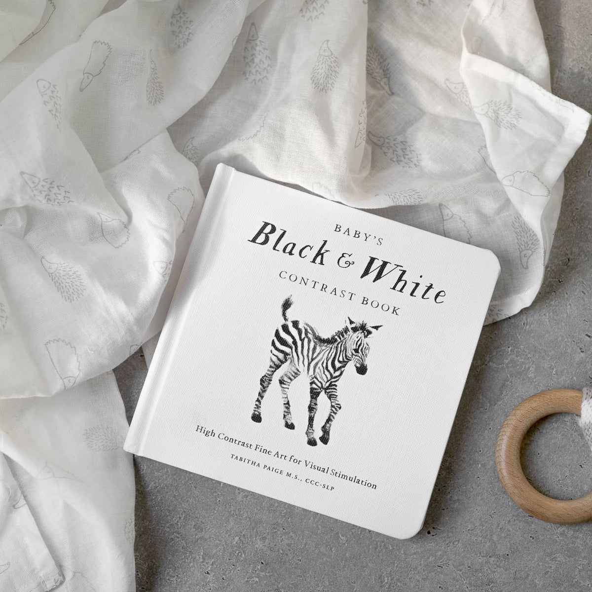 A baby contrast book titled 'Black & White Contrast Book' with a zebra illustration on the cover, styled on a gray surface with white fabric and a wooden ring toy nearby.