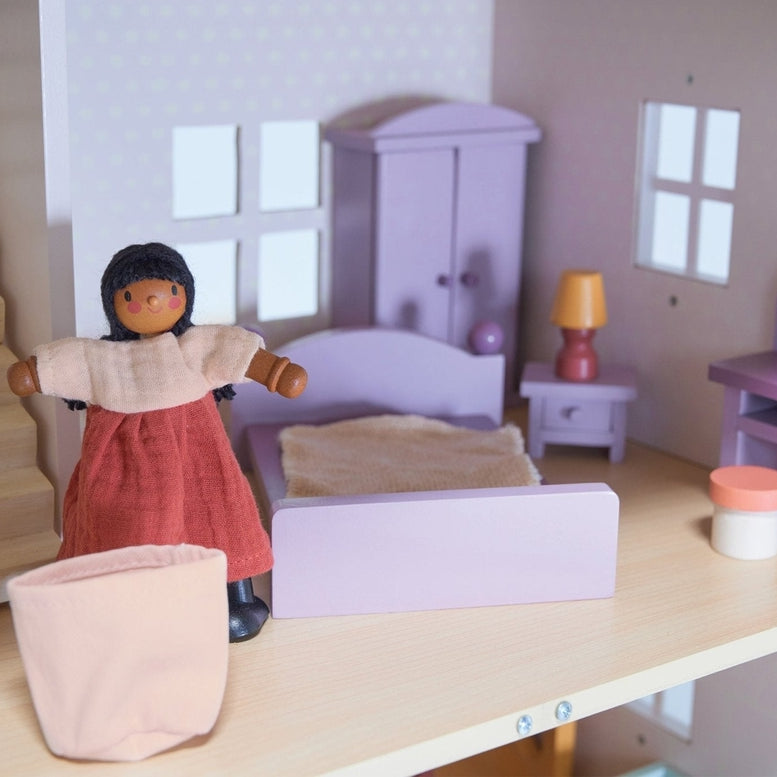 A dollhouse bedroom scene featuring a wooden doll in a reddish-brown dress standing near a white toy laundry basket in the foreground. In the background, a double bed with a purple frame and bedding sits against the wall, alongside a matching purple wardrobe, a bedside table with a yellow-shaded lamp, and a dressing table with a stool near a window.
