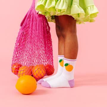 Child wearing colorful socks with fruit design, standing next to oranges and a pink mesh bag on a pink background
