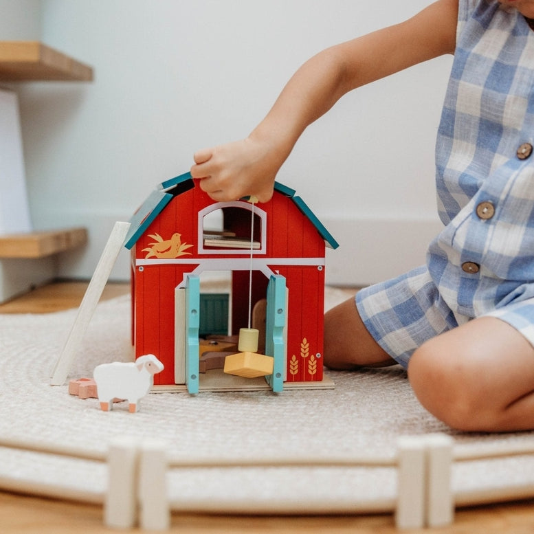 A child’s hands operating the working pulley system on the Wooden Big Red Barn Playset by Mentari, lifting a yellow magnetic hay bale toward the loft while open double doors reveal a glimpse inside the red barn.