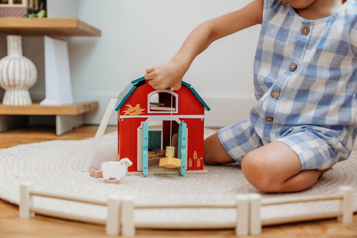 A child’s hands operating the working pulley system on the Wooden Big Red Barn Playset by Mentari, lifting a yellow magnetic hay bale toward the loft while open double doors reveal a glimpse inside the red barn.