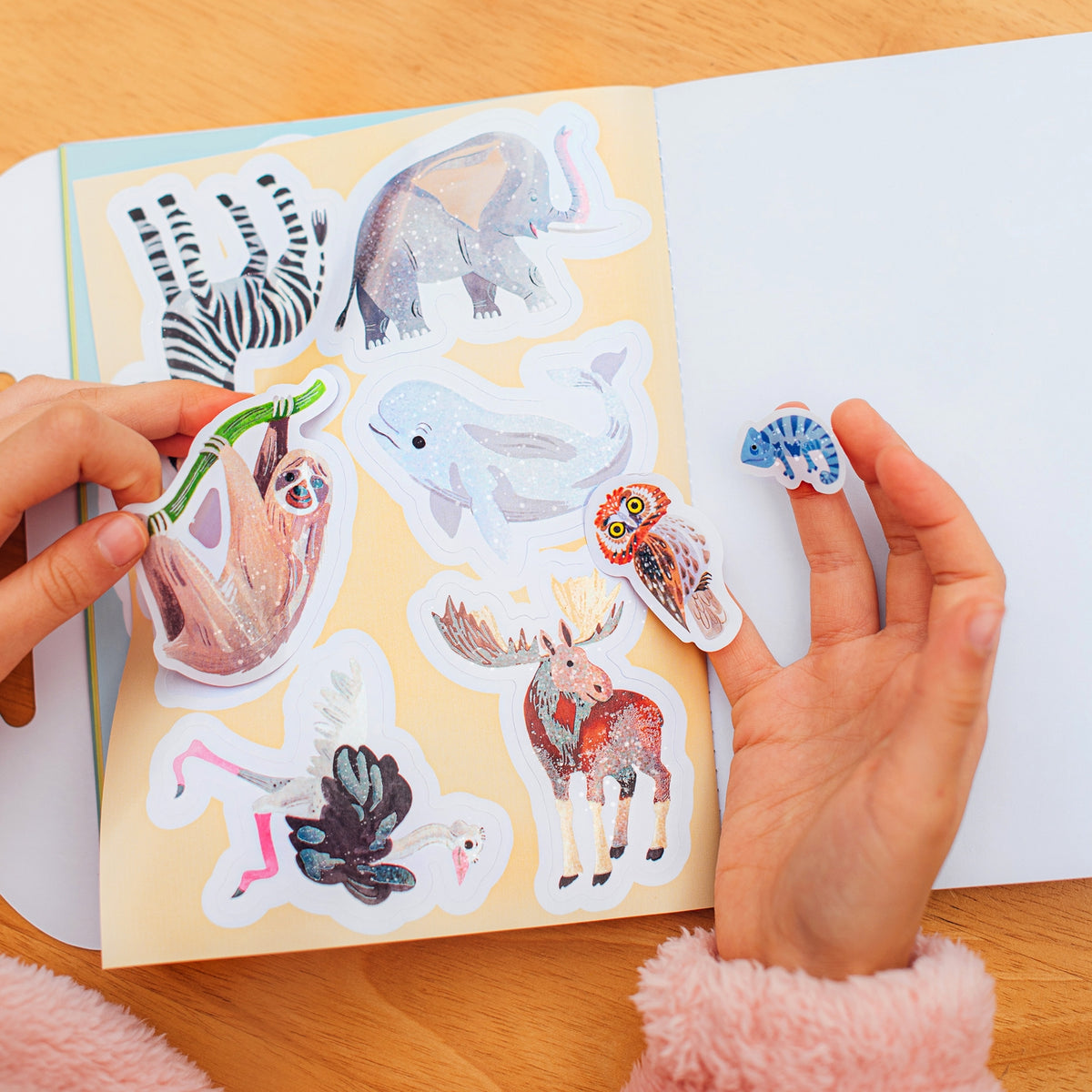 Hands holding animal stickers above a sheet of additional stickers on a wooden surface