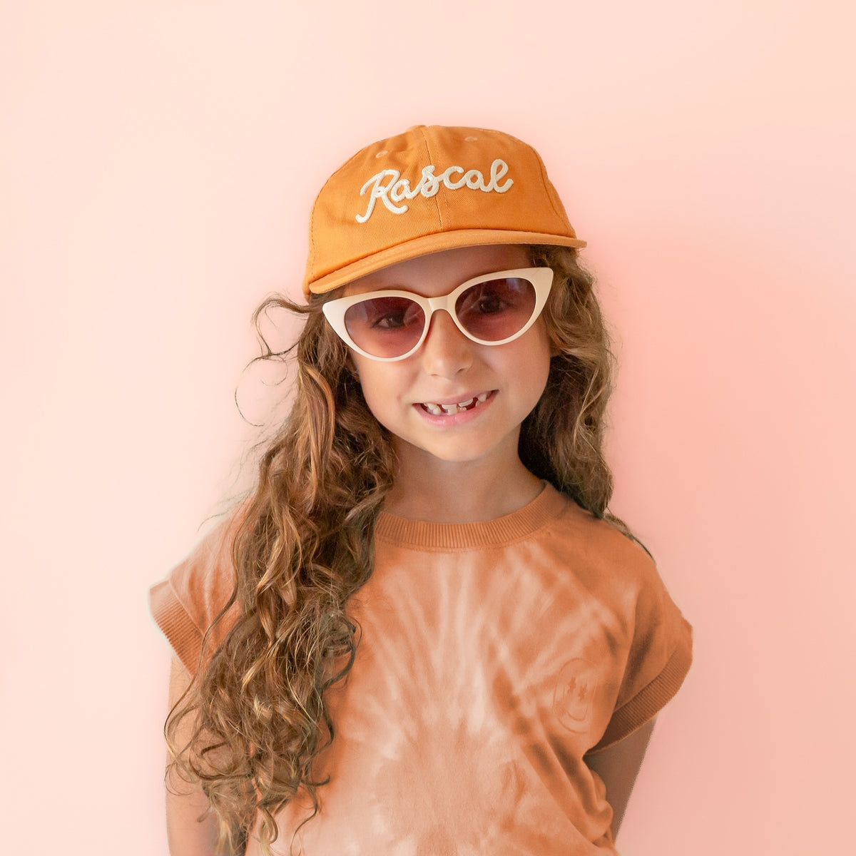 A young girl with long curly brown hair is shown from the chest up against a soft pink background. She is wearing an orange baseball cap with the word 'Rascal' embroidered in white script, white cat-eye sunglasses, and a peach tie-dye t-shirt.