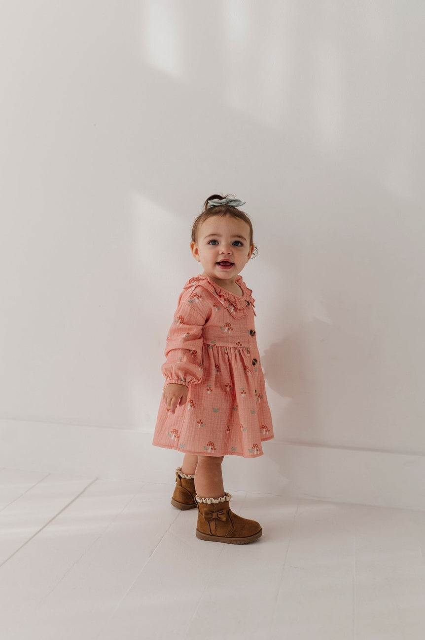 A toddler girl wearing a dusty pink long-sleeve dress with a small animal print, styled with brown suede boots, standing against a bright white wall.
