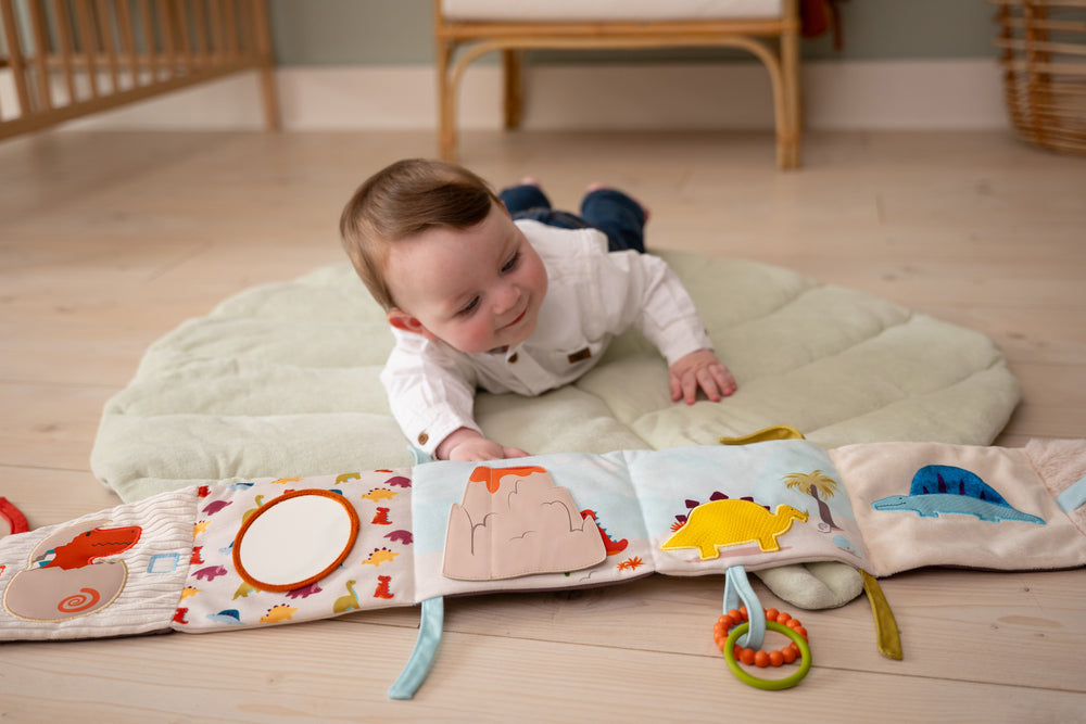 Baby playing with a soft book on a mat in a room with wooden flooring and furniture.