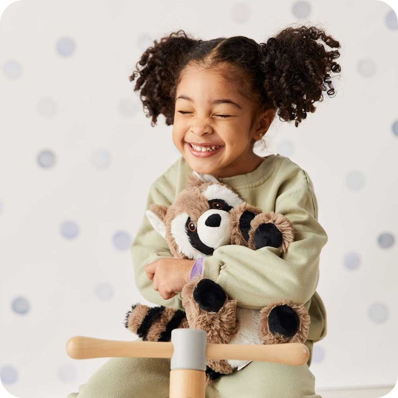 A smiling young girl with pigtails hugging a plush Raccoon by Warmies while wearing a green sweatshirt, sitting against a polka-dot background with a wooden toy in the foreground.