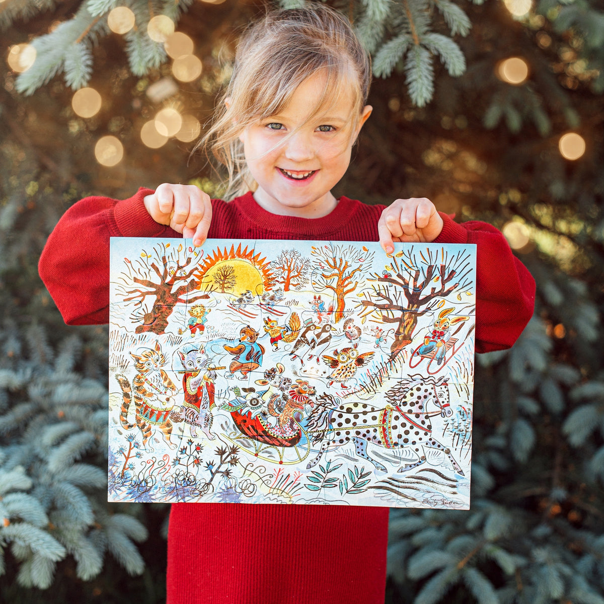 Child holding a colorful illustration of animals in a snowy scene, standing in front of Christmas trees with lights.