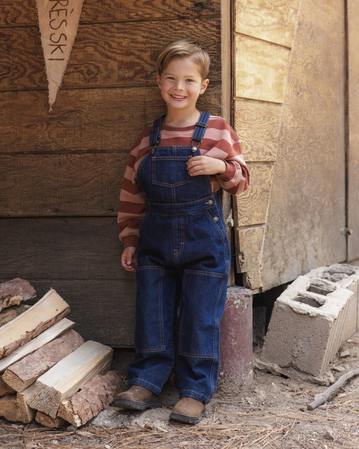 A young boy smiling and wearing blue denim overalls over a red and white striped long-sleeve shirt, standing outdoors in front of a rustic wooden wall with wood logs and a cinderblock nearby.