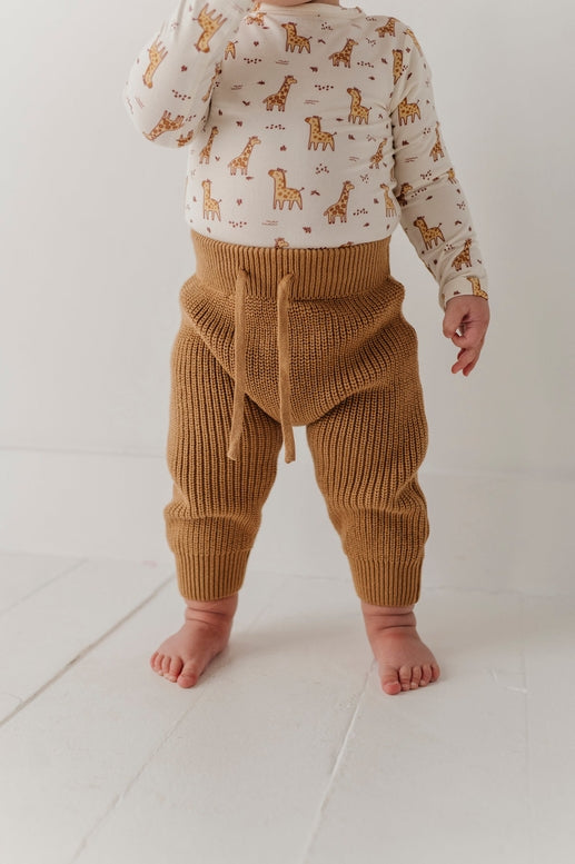 A baby standing barefoot wearing a cream long-sleeve shirt with a brown giraffe print and ribbed tan knit pants with a drawstring waist, photographed against a white studio background.