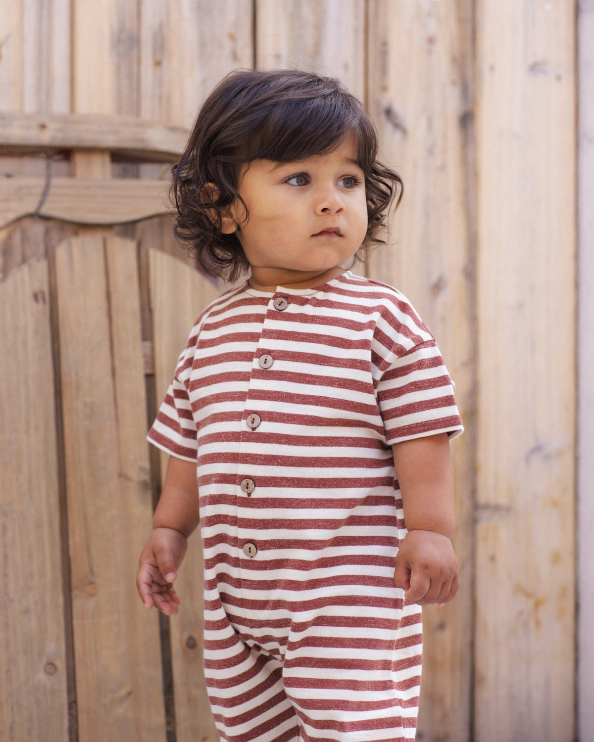 A toddler wearing a short-sleeve striped romper with brown buttons, standing in front of a light wood fence.