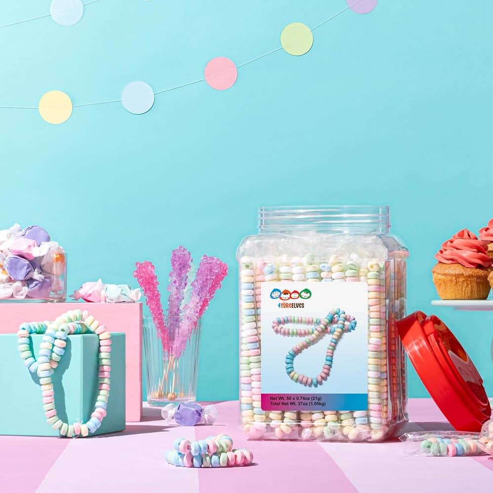 A clear plastic jar filled with pastel rainbow spiral-shaped candies stands on a pink surface against a bright turquoise background. A red lid rests beside the jar, and the scene includes decorative party props like paper garlands, a gift box with a candy necklace, pink rock candy, and cupcakes.