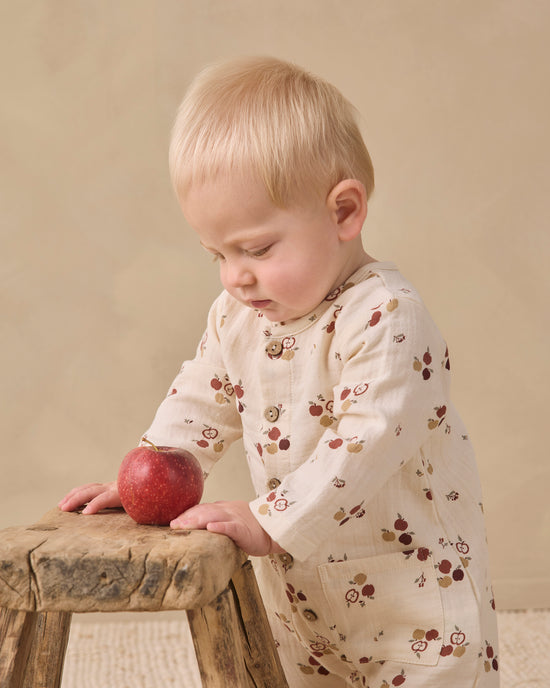 A baby wearing a cream-colored cotton pajama set with a small red and brown fruit print, leaning on a rustic wooden stool next to a red apple.