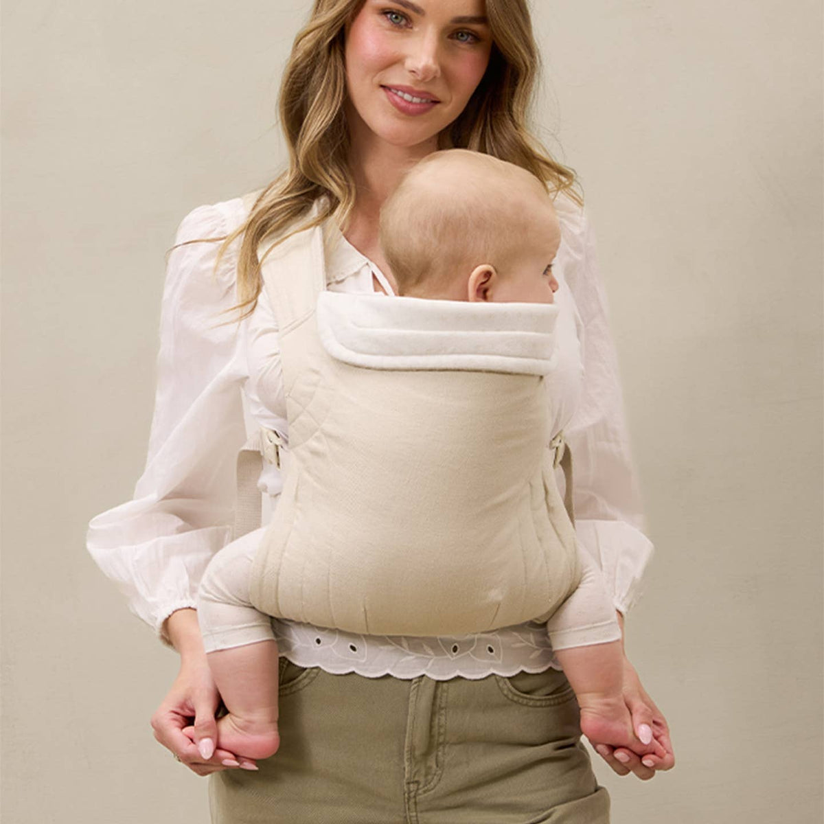 Woman holding a baby in a white baby carrier against a beige background