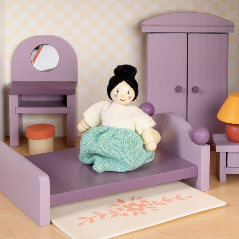 A dollhouse bedroom scene featuring a wooden doll sitting on a double bed with a purple frame. Next to the bed is a bedside table with a yellow-shaded lamp, a wardrobe against the wall, a dressing table with a mirror and stool, and a small floral rug on the floor.