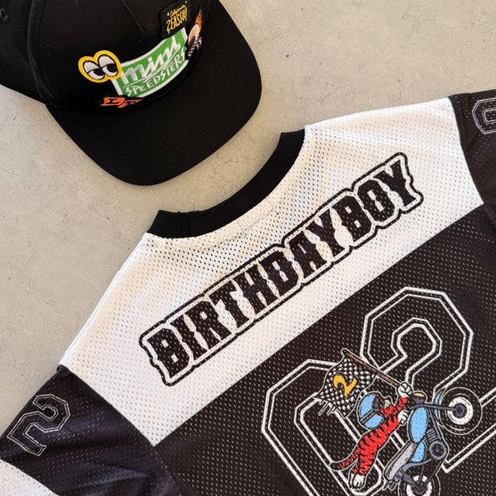 A black and white mesh baseball-style jersey and matching black cap laid flat on a light concrete surface. The cap has a colorful 'Happy Birthday' graphic, and the jersey features 'BIRTHDAYBOY' across the back with a large number 2 and a cartoon racer graphic.