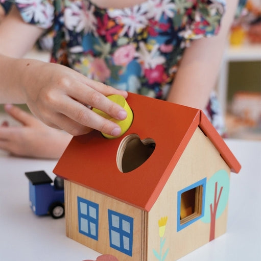 A young child looks down intently as they play with a wooden shape sorter house on a table, with an adult or older child leaning in beside them to watch. Colorful wooden blocks are scattered on the surface near the toy.