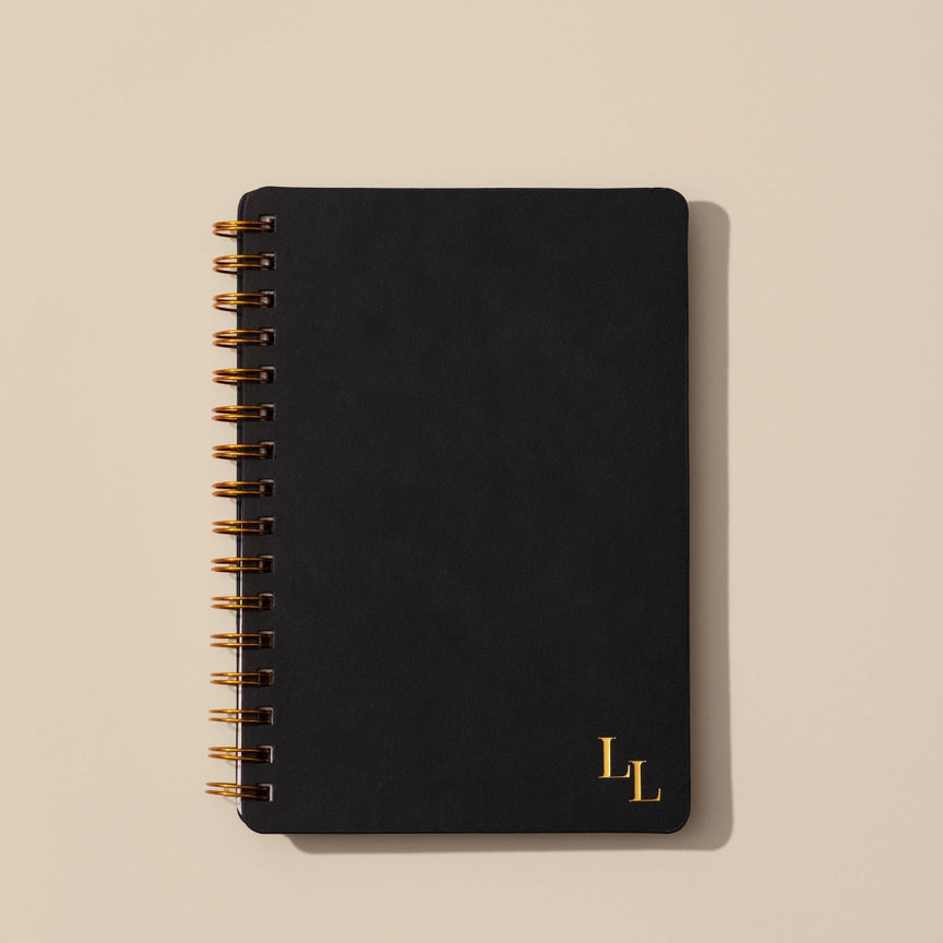 Black notebook with gold spiral binding and initials on a beige background