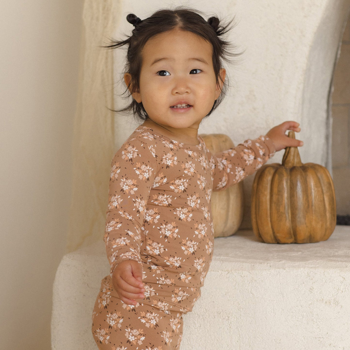 A toddler girl wearing a long-sleeve pajama set in a warm tan color with a delicate white floral pattern, standing next to a small decorative pumpkin on a textured white stone shelf.