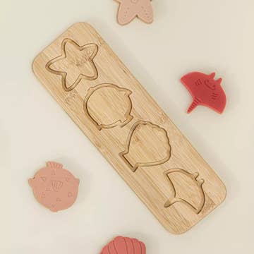 A wooden four-piece puzzle board with engraved star, circle, shell, and fish shapes, surrounded by matching pink and red plastic puzzle pieces on a light cream background.