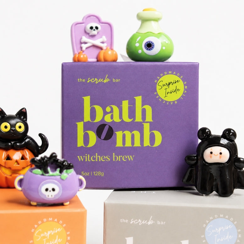 The Scrub Bar 'Witches Brew' bath bomb box in purple, styled with Halloween-themed figurines including a black cat, a green eye bottle, and a black bat figure on a clean white background.