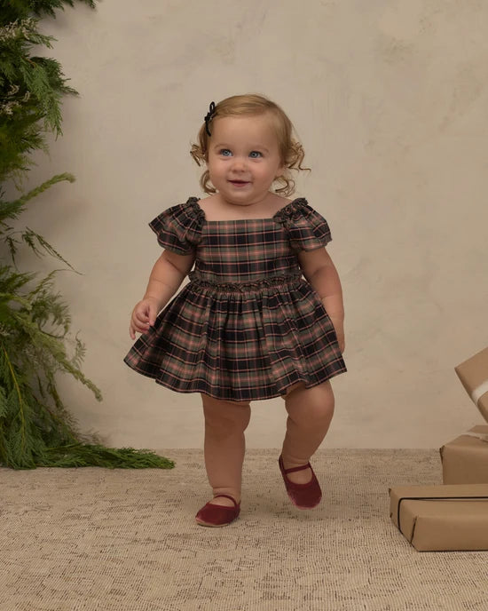 A toddler girl wearing a brown and black plaid dress with puff sleeves and maroon shoes, standing in a studio setting with a textured beige backdrop, greenery on the left, and wrapped gifts on the right.