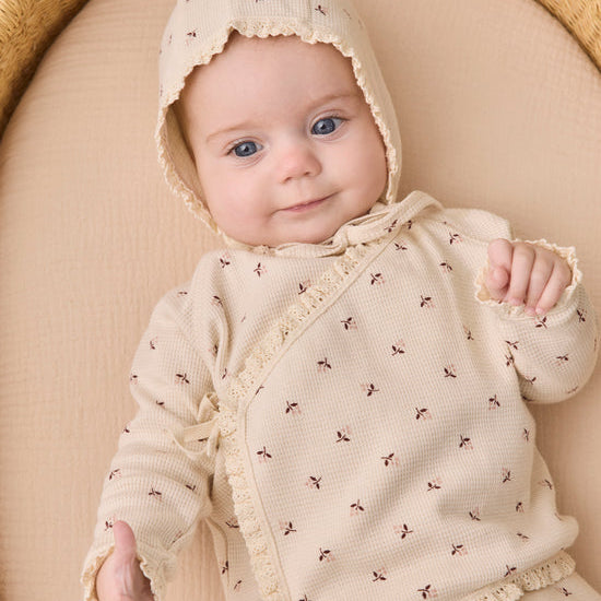 A baby wearing a cream-colored knit romper and matching bonnet with small brown embroidered motifs, lying on a beige cushion inside a woven basket.