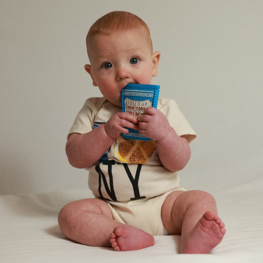 A baby sitting on a white surface against a light beige background, wearing a cream-colored short-sleeve bodysuit with black lettering and holding a blue cereal box.