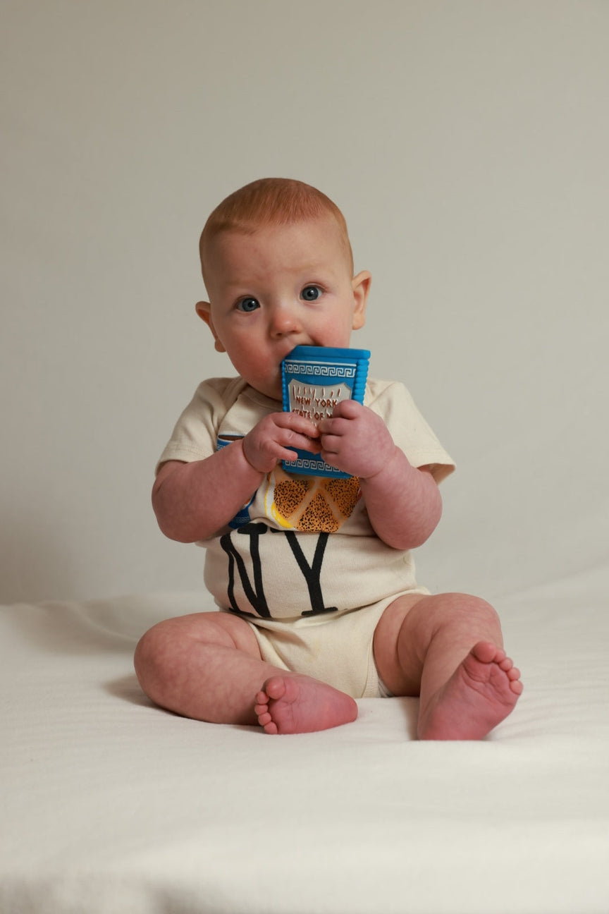 A baby sitting on a white surface against a light beige background, wearing a cream-colored short-sleeve bodysuit with black lettering and holding a blue cereal box.