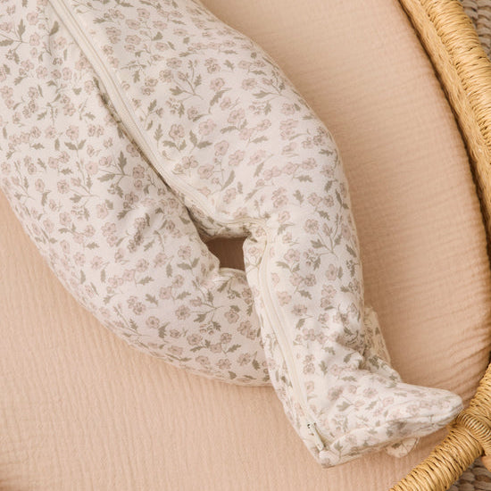 A close-up of a footed baby sleeper with a delicate beige and white floral pattern, resting on a peach-colored muslin mattress inside a woven rattan bassinet.