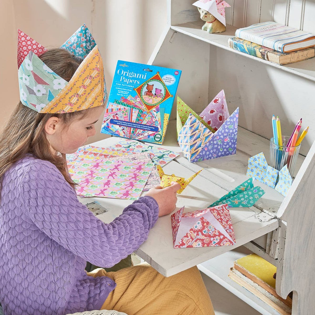 Child wearing a colorful paper crown, sitting at a table with craft materials and books.