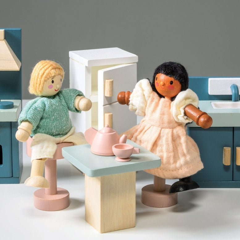 Wooden kitchen furniture set by Mentari against a white background, featuring two wooden dolls interacting at a light wood-colored table set with a teapot and two cups, surrounded by a blue stove with an oven and hood, a white refrigerator with wooden handles, and a blue sink unit with a cupboard.