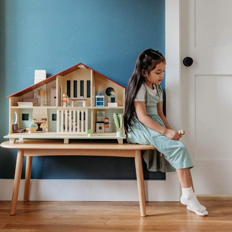 A young child sitting on a white bench by a teal wall plays with the Wooden Cedar Chalet Modernist Dollhouse by Mentari, which is arranged with miniature furniture and figures inside its open-front mid-century modern home.