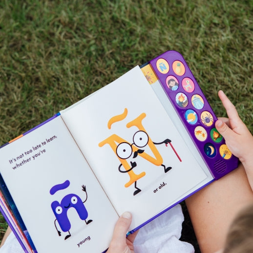 A child holding open "What Does Ñ Sound Like?: A Spanish-English Sound Book," showing an interior illustrated page featuring two animated letter characters and the text "It's not too late to learn whether you're young... or old," with a vertical panel of sound buttons on the right edge.