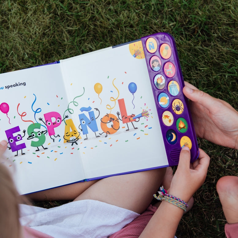A child and adult holding open "What Does Ñ Sound Like?: A Spanish-English Sound Book," showing an interior colorful spread with animated letters spelling "ESPAÑOL," balloons, and a vertical panel of interactive sound buttons on the right edge.